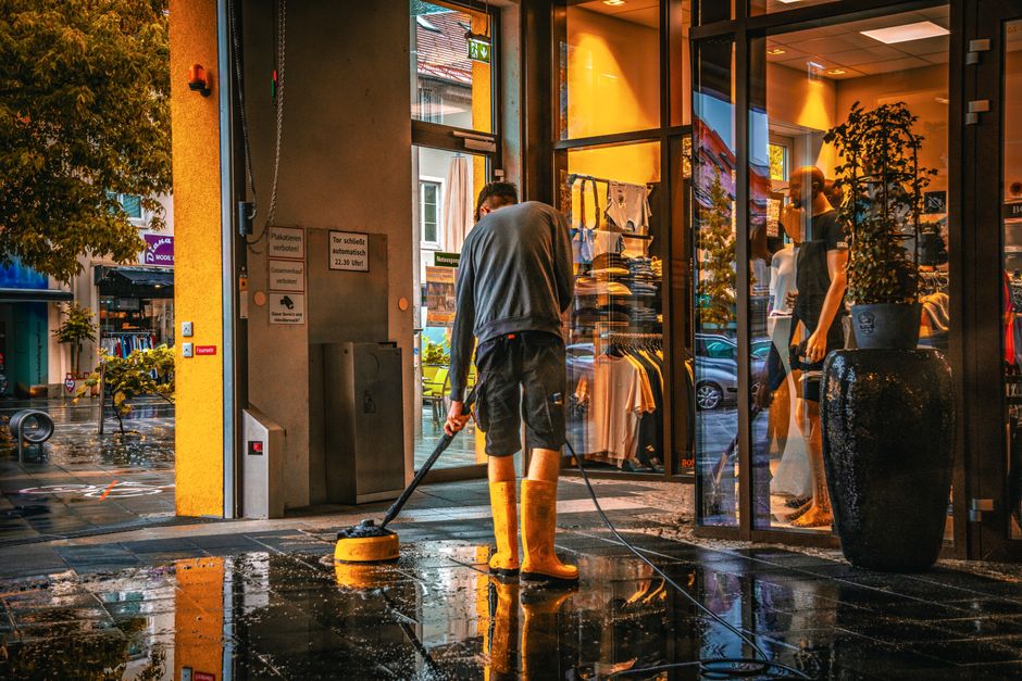A person operating a yellow pressure washer outside the entrance of a retail store in Clapham during rainy weather. The individual is wearing yellow rubber boots, black shorts, and a grey long-sleeve shirt, focused on cleaning the wet, paved surface. Inside the store, visible through large glass windows, are mannequins dressed in casual clothing, racks of folded clothes, and potted plants, with warm interior lighting illuminating the space. The exterior scene includes a tree on the left, displaying autumn foliage, and a street with parked cars and other shops in the background. Clapham Carpet Cleaning offers professional cleaning services that include surface cleaning and deep cleaning for commercial and residential properties, supporting hygiene and maintenance needs in the area.