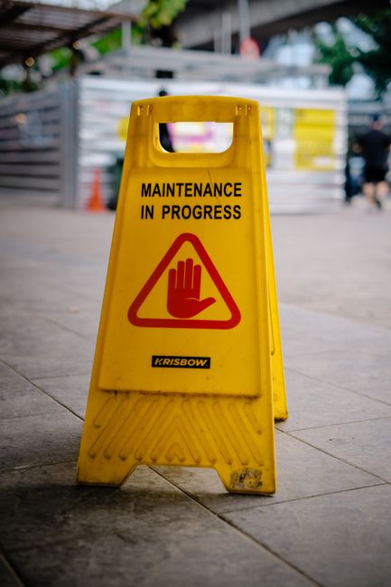 A yellow maintenance warning sign placed on a polished concrete floor outdoors, displaying the message 'Maintenance in Progress' with a red hand symbol for caution. The sign is situated in front of a background featuring metal fencing, construction materials, and green foliage, suggesting an area undergoing maintenance or cleaning. The scene is well-lit with natural daylight, and the sign appears clean and visibly prominent. Clapham Carpet Cleaning emphasizes the importance of surface hygiene and routine maintenance in both residential and commercial spaces, ensuring thorough sanitisation and deep cleaning to maintain a hygienic environment.