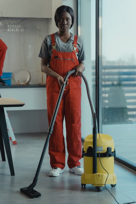 A person operating a yellow pressure washer outside the entrance of a retail store in Clapham during rainy weather. The individual is wearing yellow rubber boots, black shorts, and a grey long-sleeve shirt, focused on cleaning the wet, paved surface. Inside the store, visible through large glass windows, are mannequins dressed in casual clothing, racks of folded clothes, and potted plants, with warm interior lighting illuminating the space. The exterior scene includes a tree on the left, displaying autumn foliage, and a street with parked cars and other shops in the background. Clapham Carpet Cleaning offers professional cleaning services that include surface cleaning and deep cleaning for commercial and residential properties, supporting hygiene and maintenance needs in the area.