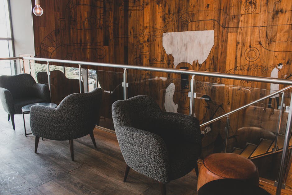 Interior area of a cafe or bar on Northcote Road featuring a wooden wall with artistic carvings and abstract designs. Four upholstered armchairs in dark and patterned fabrics are arranged in a row along a glass and metal railing. The chairs are positioned on a wooden floor, with natural light illuminating the space. In the background, a small round leather stool is visible near the stairs, suggesting a cozy seating arrangement. The scene appears clean and well-maintained, emphasizing surface cleanliness and the importance of routine surface cleaning and hygiene in hospitality environments, as promoted by Clapham Carpet Cleaning.