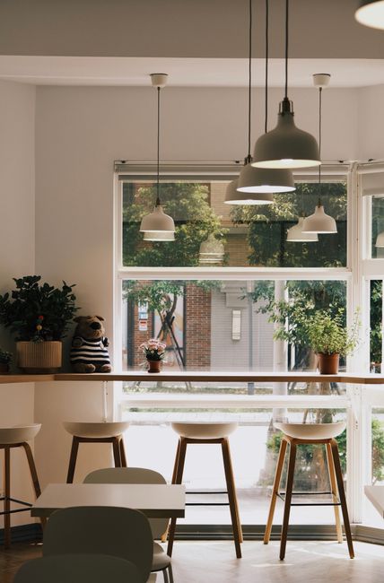 Interior view of a modern café featuring large front-facing windows allowing natural light to illuminate the space. The windows have white frames and a view of trees and brick buildings outside. Inside, there are four hanging dome pendant lights in black and white, evenly spaced above a wooden counter or bar area with three white and wooden stools. On the ledge behind the stools, potted plants and decorative items, including a small stuffed bear, add a cozy touch. The walls and ceiling are painted white, and the overall ambiance is clean and minimalist, reflecting careful surface cleaning and maintenance by Clapham Carpet Cleaning, suited for a professional hygiene and surface maintenance setting.