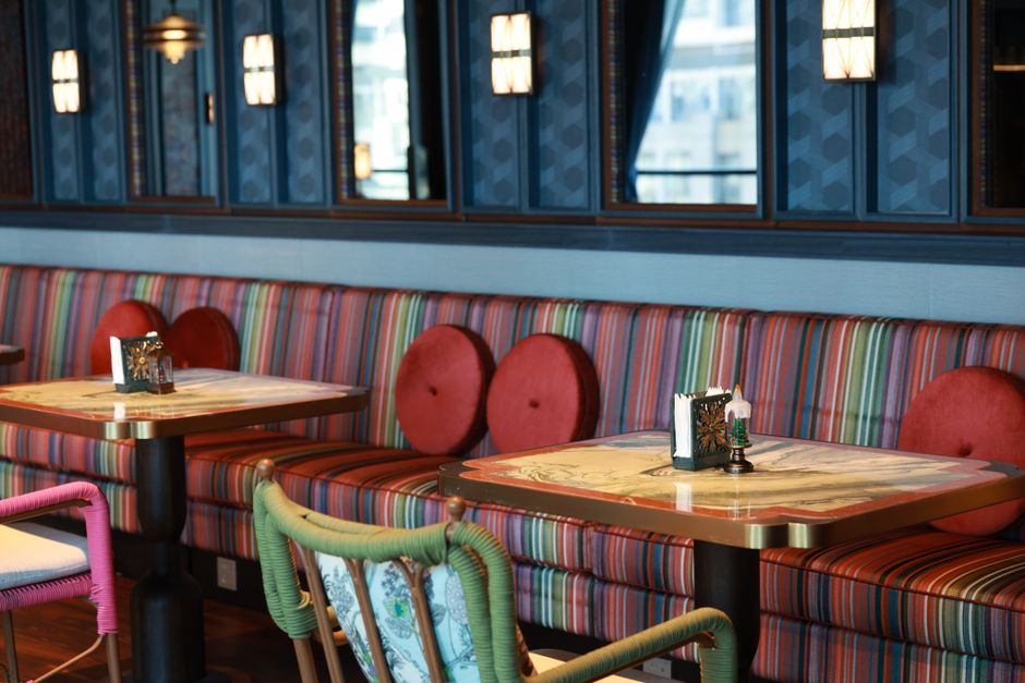 Interior area of a cafe or bar on Northcote Road featuring a wooden wall with artistic carvings and abstract designs. Four upholstered armchairs in dark and patterned fabrics are arranged in a row along a glass and metal railing. The chairs are positioned on a wooden floor, with natural light illuminating the space. In the background, a small round leather stool is visible near the stairs, suggesting a cozy seating arrangement. The scene appears clean and well-maintained, emphasizing surface cleanliness and the importance of routine surface cleaning and hygiene in hospitality environments, as promoted by Clapham Carpet Cleaning.