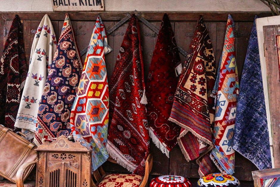 A display of several colorful, patterned rugs hanging from a wooden wall in a room, with intricate designs in red, blue, white, and other bright colors. Some rugs drape over a rail, showcasing detailed geometric and floral motifs. In front of the rugs, there is vintage wooden furniture, including a small decorative cabinet and a cushioned stool with a floral pattern. The room features soft lighting that highlights the rich textures and vibrant colors of the rugs, emphasizing the delicate craftsmanship and the cleanliness of the display area, which is part of a setting that suggests a traditional or boutique store environment, possibly showcasing handmade or collectible rugs.