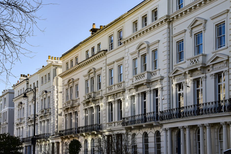 An exterior view of a row of Victorian-style terraced flats with white facades, decorative architectural details, and black wrought iron balconies. The buildings are illuminated by natural daylight, with some shadows cast by nearby trees. The street features a classic black lamppost and small trees lining the pavement. This image reflects the elegant and historic architecture typical of Clapham SW4, presented in a clean and well-maintained setting, suitable for surface cleaning and deep cleaning services by Clapham Carpet Cleaning, focusing on maintaining the hygiene and visual appeal of residential exteriors in Victorian flats.