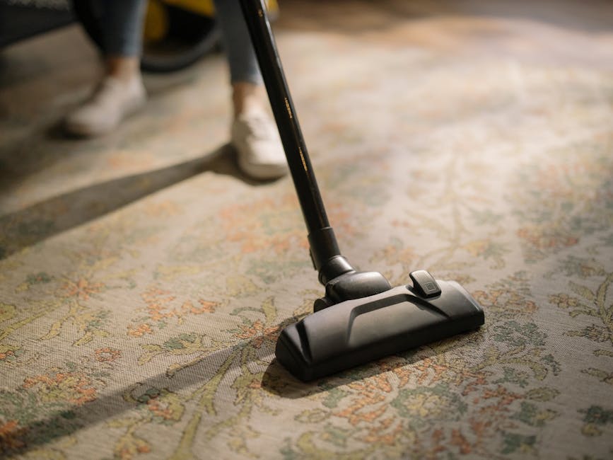 Close-up image of a vacuum cleaner nozzle actively cleaning a patterned area rug in a residential setting. The rug features intricate floral motifs in shades of cream, light green, and muted orange. The vacuum cleaner, with a black body and a control button, is positioned on the edge of the rug, with a person's white sneakers and part of their grey pants visible in the background, indicating active cleaning in a living room or similar space. The area appears well-maintained and clean, with natural lighting highlighting the surface's detail, emphasizing surface cleaning and maintenance by Clapham Carpet Cleaning.