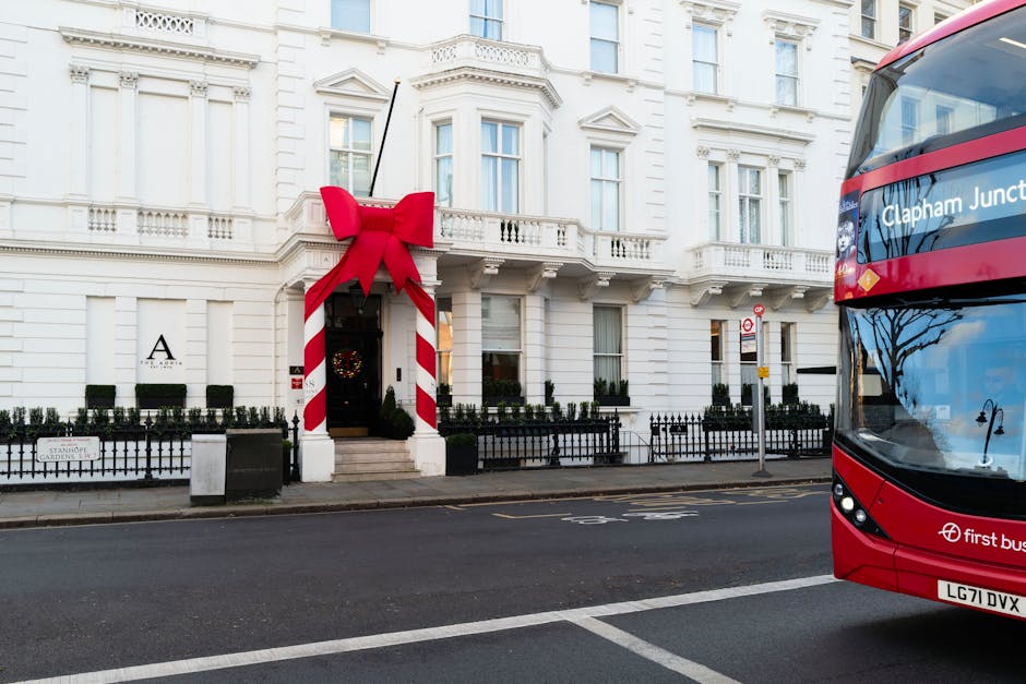 A large white residential building decorated with a giant red and white striped Christmas ribbon and bow above the entrance, which features a black door and a small wreath. The building has multiple windows with white frames and decorative molding, set behind a low black metal fence with small shrubbery along the front. The street in front is paved with dark asphalt, and a red double-decker bus is partially visible on the right side of the image. The scene is well-lit with natural daylight, highlighting the clean, pristine appearance of the building’s exterior, indicative of professional surface and building cleaning services offered by Clapham Carpet Cleaning near Clapham Junction Station.