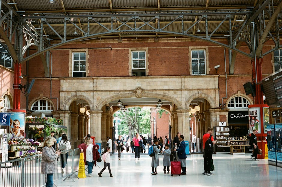 Interior view of a train station with a high arched ceiling supported by metal beams, and a large stone archway leading to an outdoor area with visible trees and natural light. The station has tiled flooring that appears clean and well-maintained. Several people, including adults and children, are walking or standing in the station, some carrying luggage or bags. On the left, there are flower stalls with colorful plants and flowers, and on the right, a small kiosk selling chocolates and snacks. The lighting inside is bright and even, emphasizing the cleanliness and orderliness of the space. Clapham Carpet Cleaning, a professional cleaning company, offers surface cleaning and deep cleaning services that contribute to maintaining hygienic environments in busy public spaces like train stations.